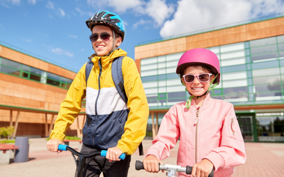 a young boy and girl in cycling helmets, standing on scooters in front of a modern school building