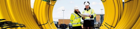 a man and woman in high vis and helmets inspect a pile of coiled plastic yellow pipes, photo taken from inside the coils looking outwards