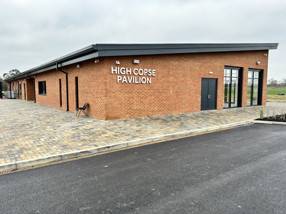 a modern sports pavilion with tall glass windows on two sides and High Copse Pavlion in stencilled metal letters along the side