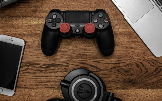 A phone, playstation controller, headphones and laptop on a table