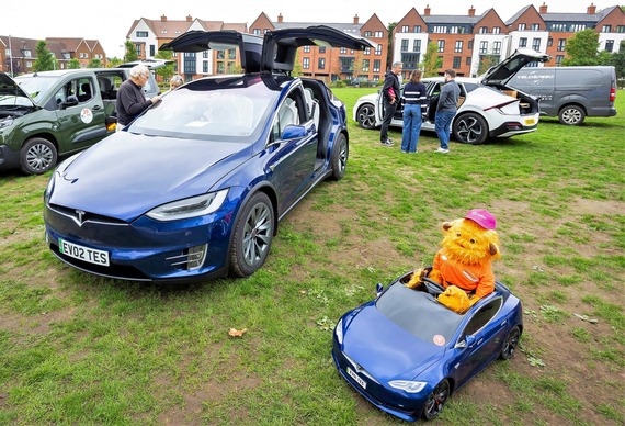 a man looking at an electric vehicle with its doors open on Elms Field, and a miniature remote control EV nearby with a furry puppet driving it