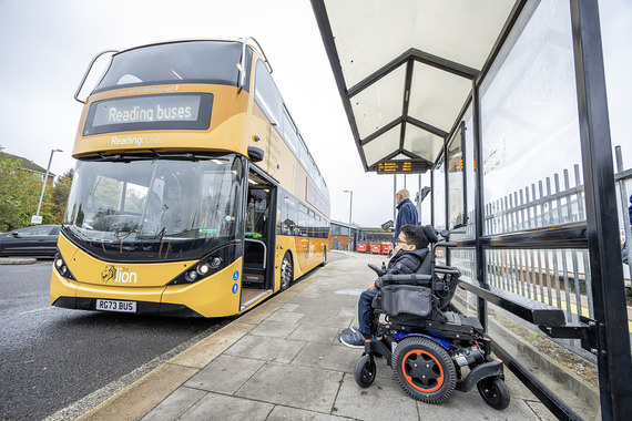 a yellow Reading Buses double-decker pulls into a covered shelter as a man in an electric wheelchair looks on and waits to board