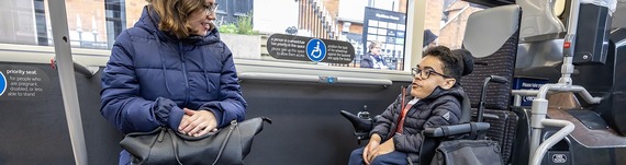 a man aboard a bus in an electric wheelchair, talking to a woman sitting opposite him