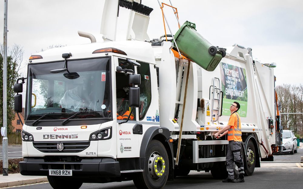 A green bin being lifted on the side of a rubbish and recycling collection lorry, overseen by a man in orange high vis