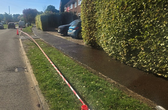 a visibly wet, newly laid footway in a leafy residential street, taped off with cones while it dries