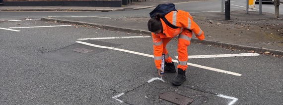 a worker in orange high vis sprays lines around a grate in the middle of the road to mark it for repair