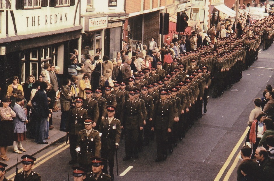 slightly faded 1978 photo of a large military parade marching past the Redan pub and a fish and chip shop in central Wokingham