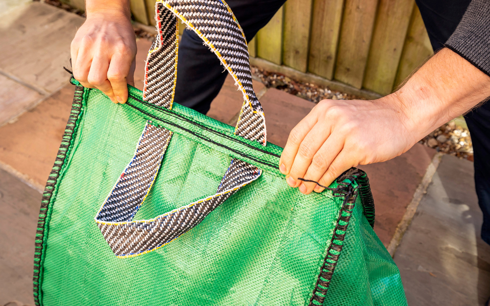 Hands grasping striped handles of a green recycling bag. A wooden fence and paved ground are in the background.