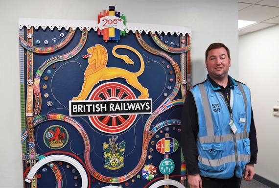 a man in SWR branded high vis stands and smiles next to a large artwork made up of painted sections of toy railway track at Wokingham station