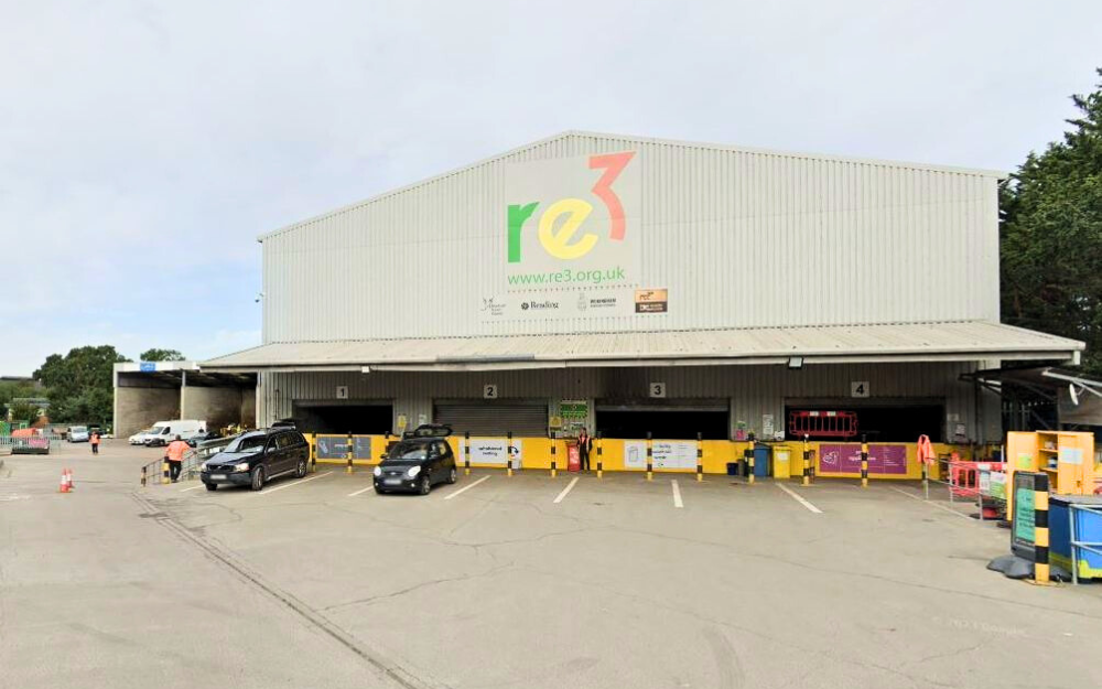Large pale grey re3 building with logo, four loading bays and yellow barriers; cars and workers in high-vis vests active in the yard.