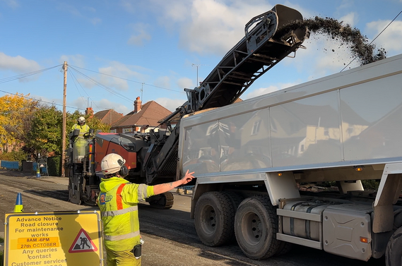 A resurfacing lorry sprays a jet of hot black asphalt into the back of a dispensing machine, guided by a man in high vis