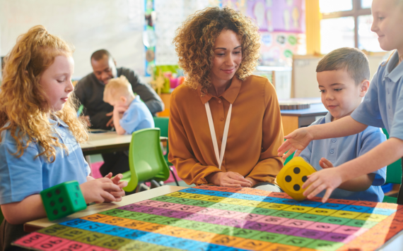 Two children in school uniform sat at a table with a teacher, playing a maths game with a large foam dice