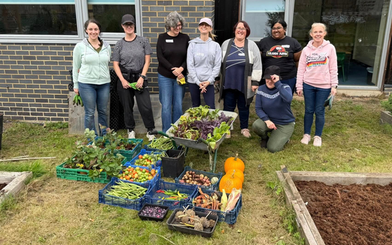 Harvest of pumpkins, squashes, beans and leafy greens displayed in crates and a wheelbarrow, with volunteers standing behind.