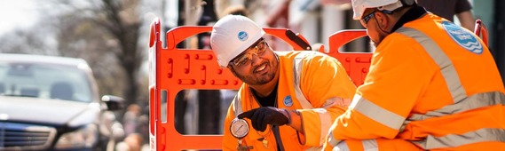 Two Thames Water workers smile at each other as they crouch by a manhole in the pavement and check water pressure