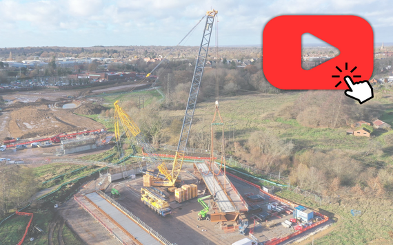 aerial shot of a huge crane carrying horizontal steel beams for a new bridge in the middle of a field, with "click to play video" graphic