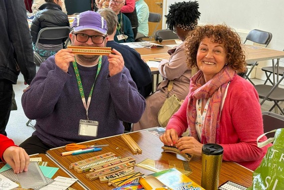 a man in a baseball cap, sitting next to a woman, smiles and holds up a toy wooden railway track which he has coloured in