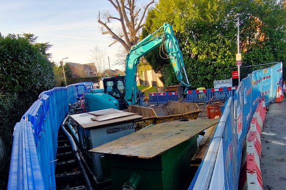 a works site behind tall fencing on Evendons Lane, with a big hole in the middle of the road and a digger working on it