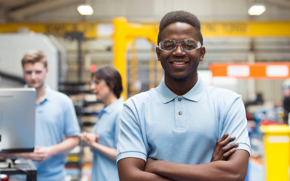 A young adult male smiling in a warehouse, wearing safety goggles