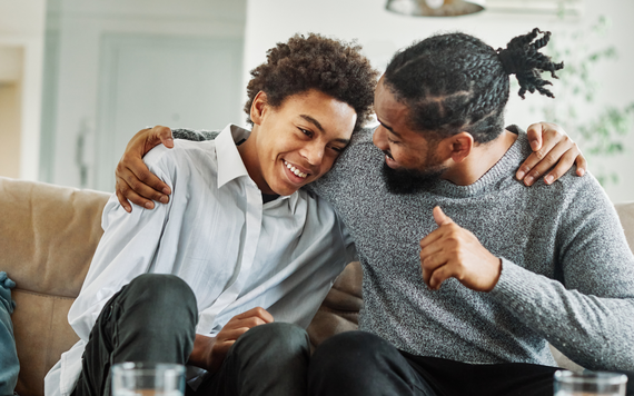 A teenage son and father sat together on a sofa, with father's arm around son