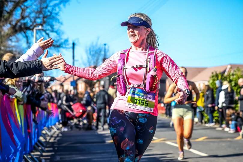 A female runner in a pink long-sleeve top and patterned leggings, wearing a hydration vest and visor, high-fives supporters along a road.