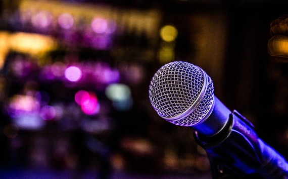 Close-up of a silver microphone with a metal mesh in the foreground. The background features colourful, out-of-focus stage lights.