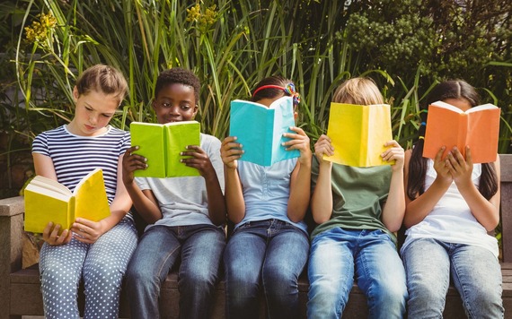Five children sitting on a wooden bench outdoors, each holding a bright book and reading. A backdrop of tall green plants provides a natural setting.
