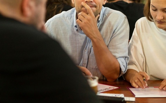 People siting at a wooden table; a man in a light blue shirt resting his chin on his hand. A woman in white to his right writes on papers.
