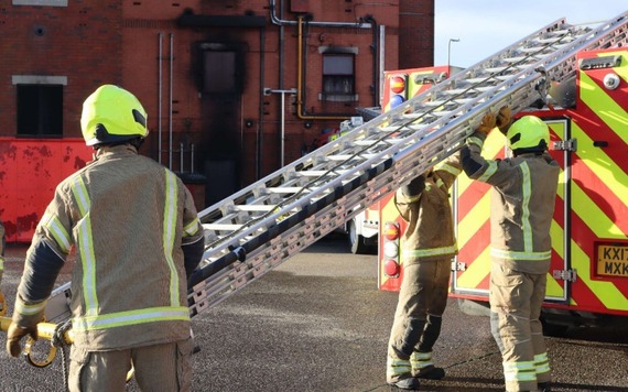 Firefighters in turnout gear and neon helmets lift an extendable ladder from a red-and-yellow fire engine. 