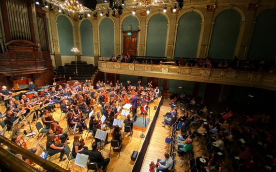 A fullorchestra performing on a wooden stage. The conductor directs from the podium as the audience sits in rows and on a balcony.
