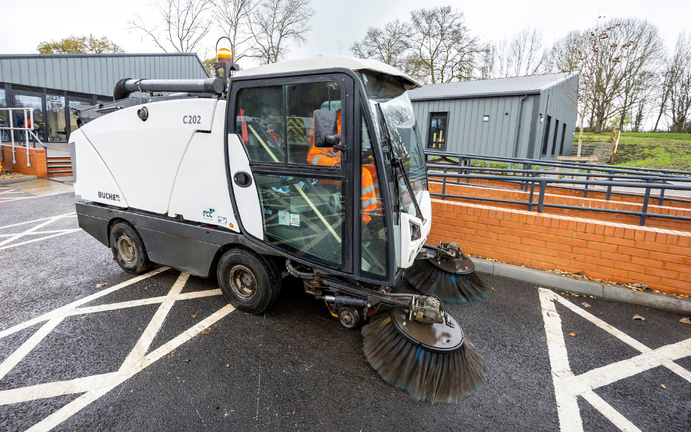 A white compact street sweeping machine being driven by a man in orange high visibility clothing.