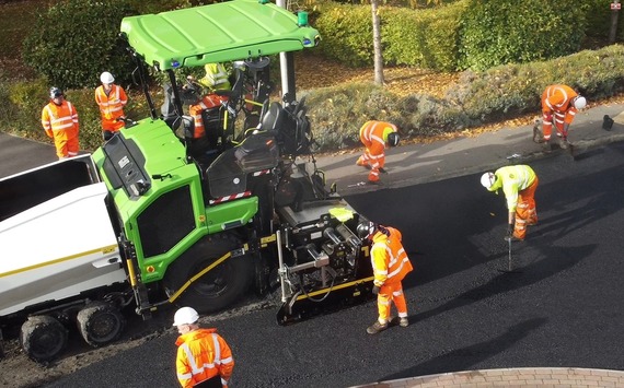 aerial view of a large machine laying asphalt at a roundabout as men in orange high viz even it out and sweep the road behind it