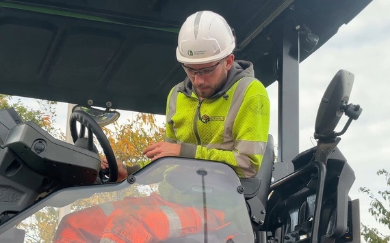 A man in high viz, with a helmet and eyewear, sitting at the wheel of a road resurfacing vehicle and looking at the road