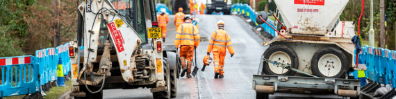 wide shot of a road resurfacing site with big vehicles either side of the road and men in orange high vis looking busy