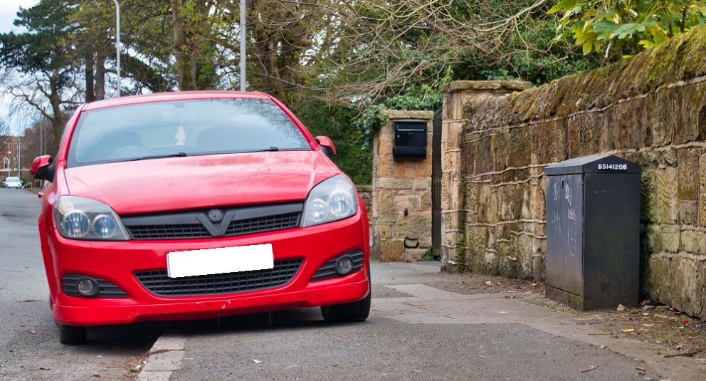 a red car parked on a pavement, not leaving enough space for anyone to pass