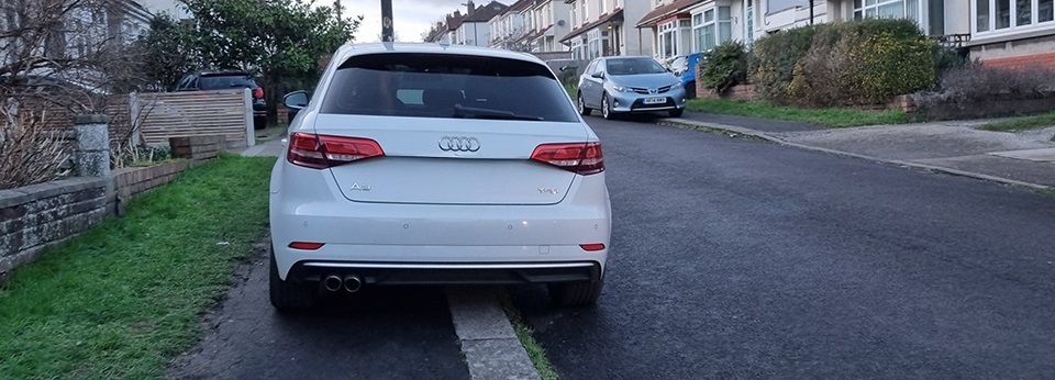 a white car parked on a pavement, not leaving enough room for anyone to get past
