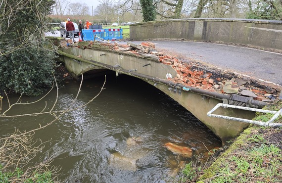a small rural bridge in Swallowfield, closed off with one side wall completely destroyed, and no barrier between the road and the river below