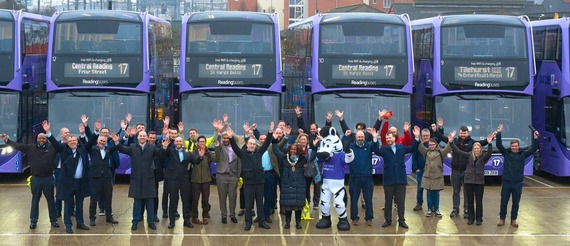 a large group of people, including one person in a cuddly zebra costume, cheer in front of a long line of purple electric buses