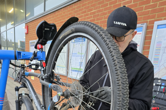 a man in a black fleece inspects a bike on a rack and adjusts its gears with a tool, outside Wokingham station