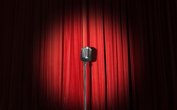 A vintage metal microphone on a stand centered before red theatre curtains on a stage.