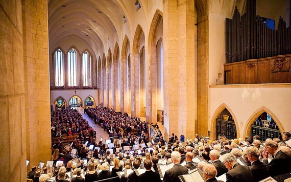 Interior of large stone church with tall arches and a vaulted ceiling, a full orchestra performs on a front stage in front of a crowd.