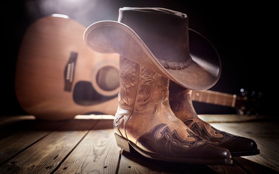 A pair of brown leather cowboy boots on a wooden floor with a brown cowboy hat resting on top. A wooden guitar in the background.