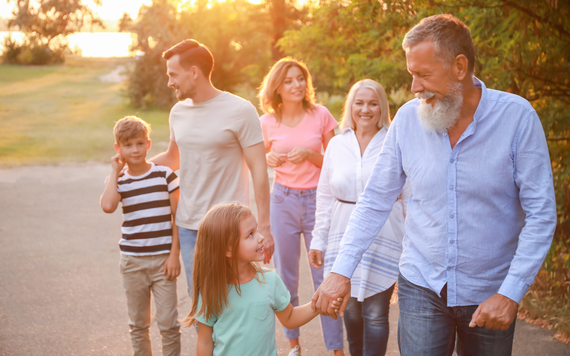 a family of adults and children walking through a green space at sunset, with a little girl in foreground holding an older man's hand