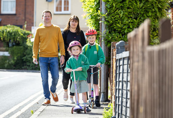 a young girl and older boy riding scooters down a sunny street with their parents behind them