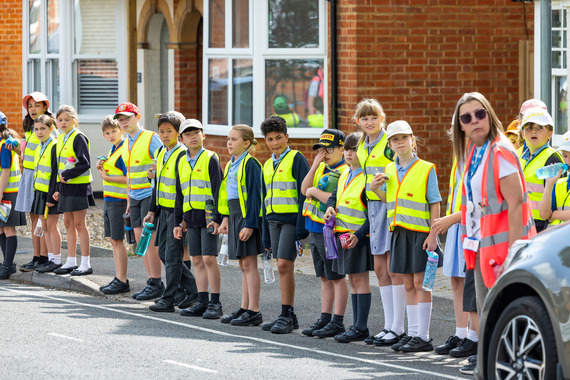 large group of children in high viz vests, looking left under an adult's supervision while preparing to cross a road