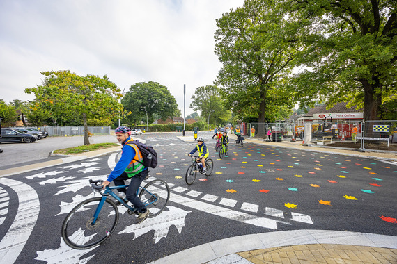 an adult cyclist rides across a surfaced junction painted with a colourful leaf pattern, with children riding behind