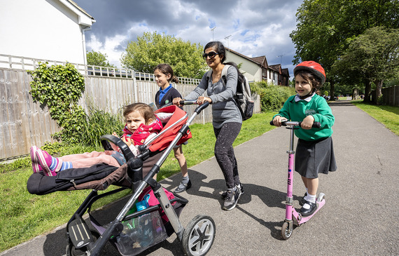 a mother with her young child in a pushchair on a tidy suburban footpath, with one daughter on a scooter and another walking alongside