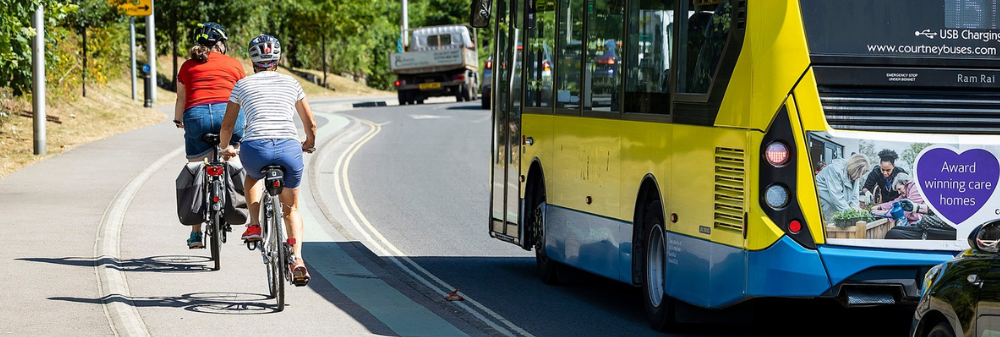 two cyclists riding together along a segregated cycle track, with a bus driving in the road alongside them
