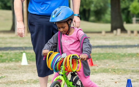 a very young girl in a pink outfit and bike helmet rides a balance bike, while looking at plastic hoops dangling from the handlebars