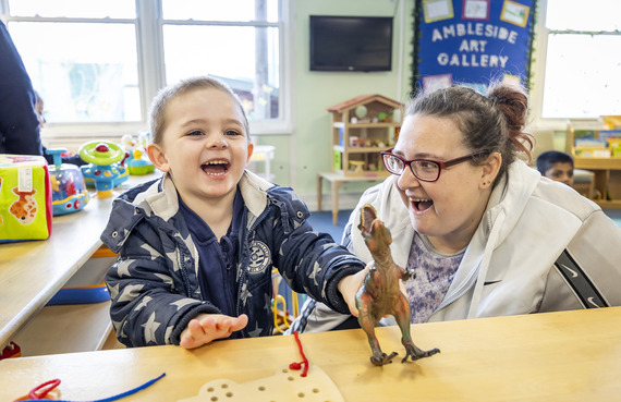 a small child sits at a table and laughs while holding up a toy dinosaur, as a woman looks on smiling