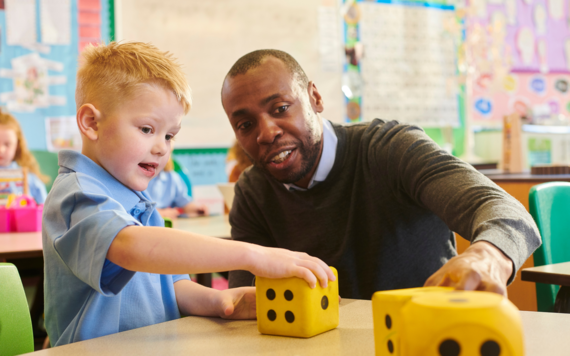 A child in school uniform sat with a teacher, rolling a large foam dice during a maths game